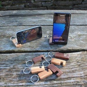 Peut inclure: Deux téléphones portables sont posés sur des supports de téléphone en bois. Les supports sont attachés à des porte-clés. Il y a six autres supports de téléphone en bois sur une table en bois.
