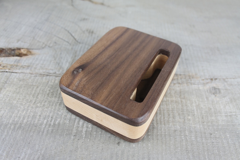 Puede incluir: Un soporte de tel&eacute;fono de madera hecho de nogal y arce. El soporte tiene un hueco para un tel&eacute;fono y un acabado de madera natural.