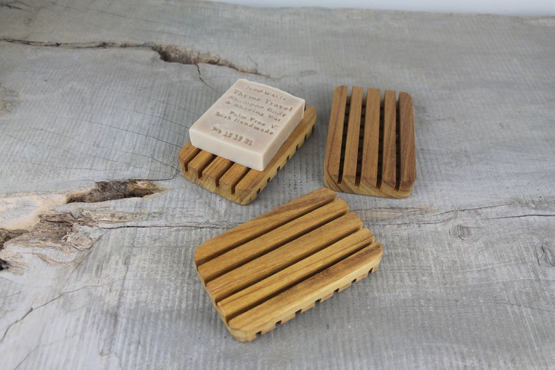 May include: Three wooden soap dishes with raised ridges. The soap dish on the left has a bar of soap on it. The soap is labeled "Thyme Travel & Mountain Mint Soap Bar, Palm Free,  Soap Handmade, Jul 15 18 21"