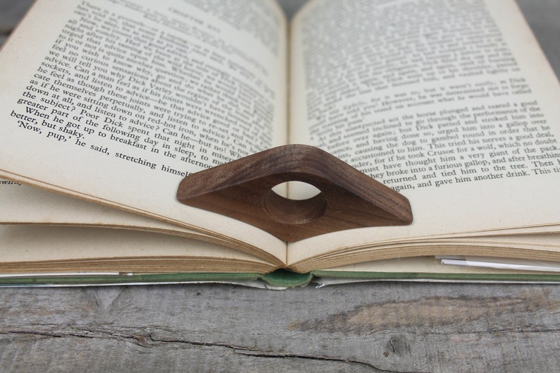 May include: A wooden book holder with a circular opening in the center. The holder is brown and is holding open a book with white pages. The book is resting on a wooden surface.