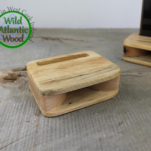 Puede incluir: Soporte de teléfono de madera con forma rectangular y una ranura para sujetar el teléfono. El soporte está hecho de madera clara y tiene un patrón de vetas natural. El logotipo de Wild Atlantic Wood es visible.