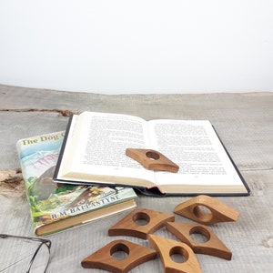 May include: Six wooden book page holders in the shape of a teardrop with a hole in the center. The holders are on a wooden surface with a book and a pair of glasses.