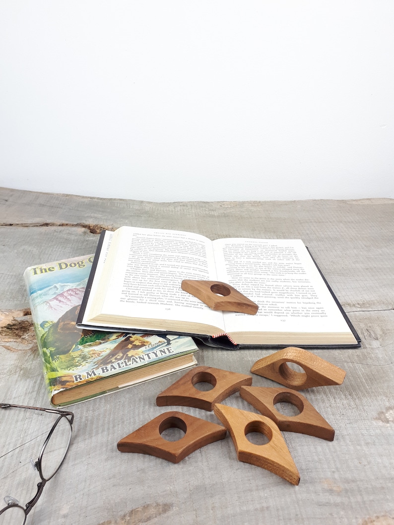 May include: Six wooden book page holders in the shape of a teardrop with a hole in the center. The holders are on a wooden surface with a book and a pair of glasses.
