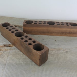 May include: Two wooden organizers with multiple round holes for storing small items. The organizers are made of dark brown wood and are sitting on a light brown wooden surface.
