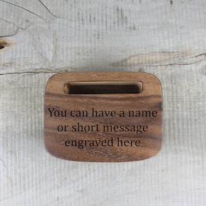 Puede incluir: Un soporte de tel&eacute;fono de madera con una ranura para un tel&eacute;fono. El soporte est&aacute; grabado con el texto "You can have a name or short message engraved here."