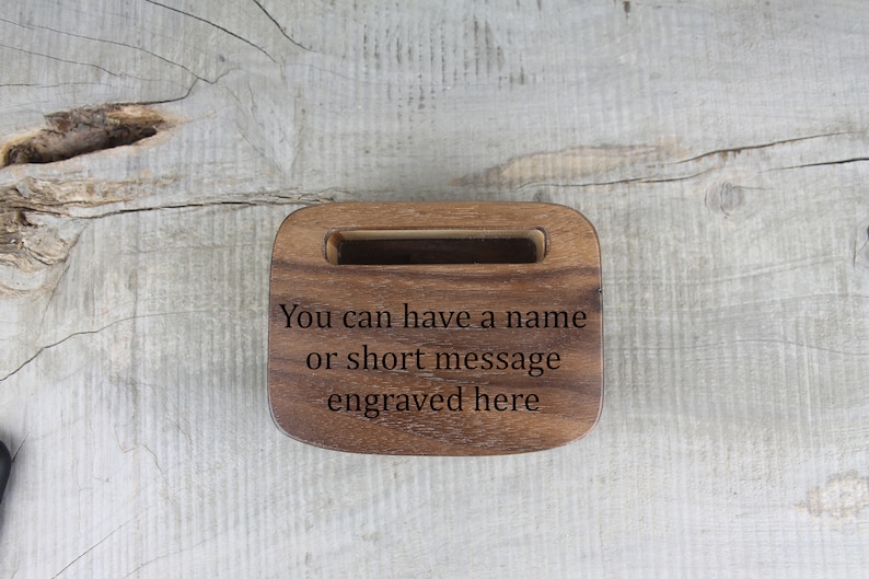 Puede incluir: Un soporte de tel&eacute;fono de madera con una ranura para un tel&eacute;fono. El soporte est&aacute; grabado con el texto "You can have a name or short message engraved here."