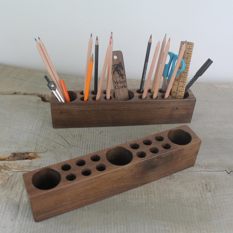 May include: Two wooden pencil holders with multiple slots for pencils, pens, and other art supplies. The top holder has a label that reads "West Cork".