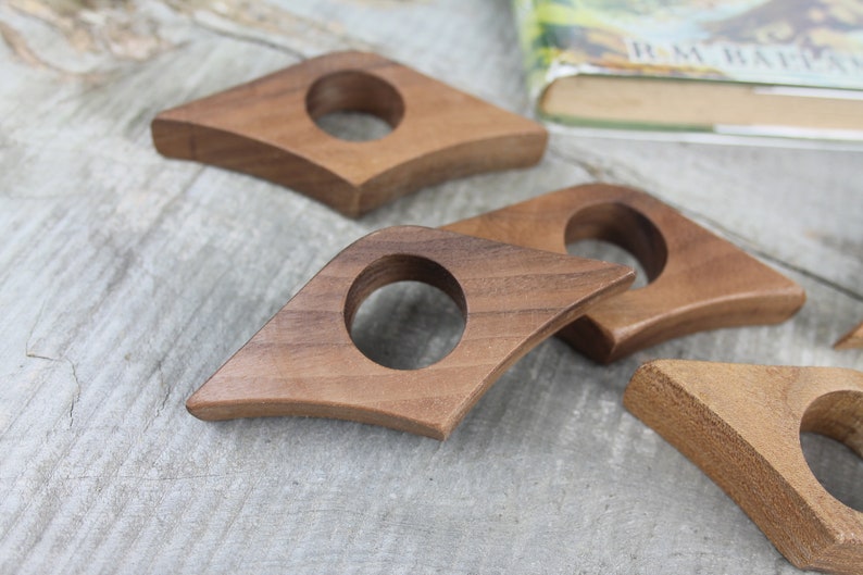 May include: Four wooden book page holders in the shape of a diamond with a rounded hole in the center. The holders are made of dark brown wood and are arranged on a light gray wooden surface.