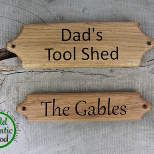 May include: Two wooden signs with rounded edges and black lettering. The top sign reads "Dad's Tool Shed". The bottom sign reads "The Gables".