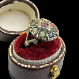 Puede incluir: Un anillo de plata con una piedra cuadrada central rodeada de piedras preciosas más pequeñas y coloridas. El anillo se exhibe en un joyero marrón forrado de terciopelo rojo con un cierre dorado. Las piedras preciosas incluyen rojo, naranja, amarillo y azul.
