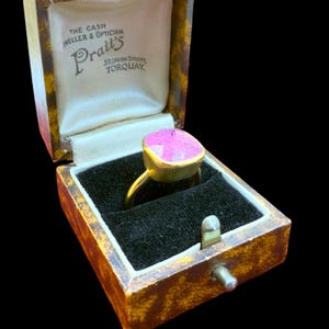 May include: A gold ring with a large, pink, square-cut gemstone, displayed in an open vintage jewelry box. The box is lined with cream-colored satin and black velvet. The box has the text "Pratt's" and "The Cash Jeweller & Optician".