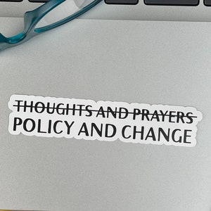 May include: A white sticker with black text on a silver laptop. The sticker says "Thoughts and Prayers" with a line through it and "Policy and Change" below it.
