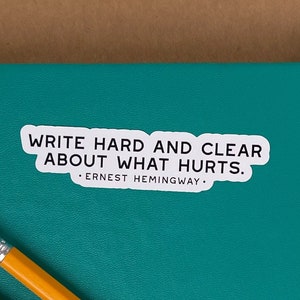 May include: A teal notebook with a white sticker that reads "Write hard and clear about what hurts. - Ernest Hemingway."