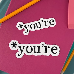 May include: Two white stickers with black text on a pink surface. The text on the stickers reads "*you're".
