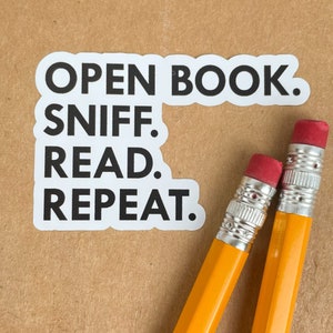 May include: White sticker with the text "OPEN BOOK. SNIFF. READ. REPEAT." on a brown background with two yellow pencils with red erasers.