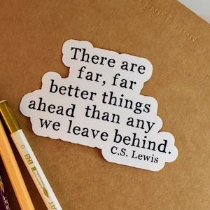 May include: A white sticker with the quote "There are far, far better things ahead than any we leave behind. C.S. Lewis" is placed on a brown notebook. Three pencils, purple, beige, and white, are positioned below the sticker.