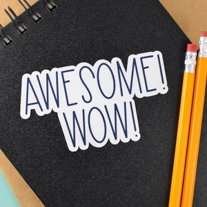 May include: A black spiral-bound notebook with a white sticker that reads "AWESOME! WOW!" in navy blue lettering. Two yellow pencils with pink erasers are positioned to the right of the notebook.