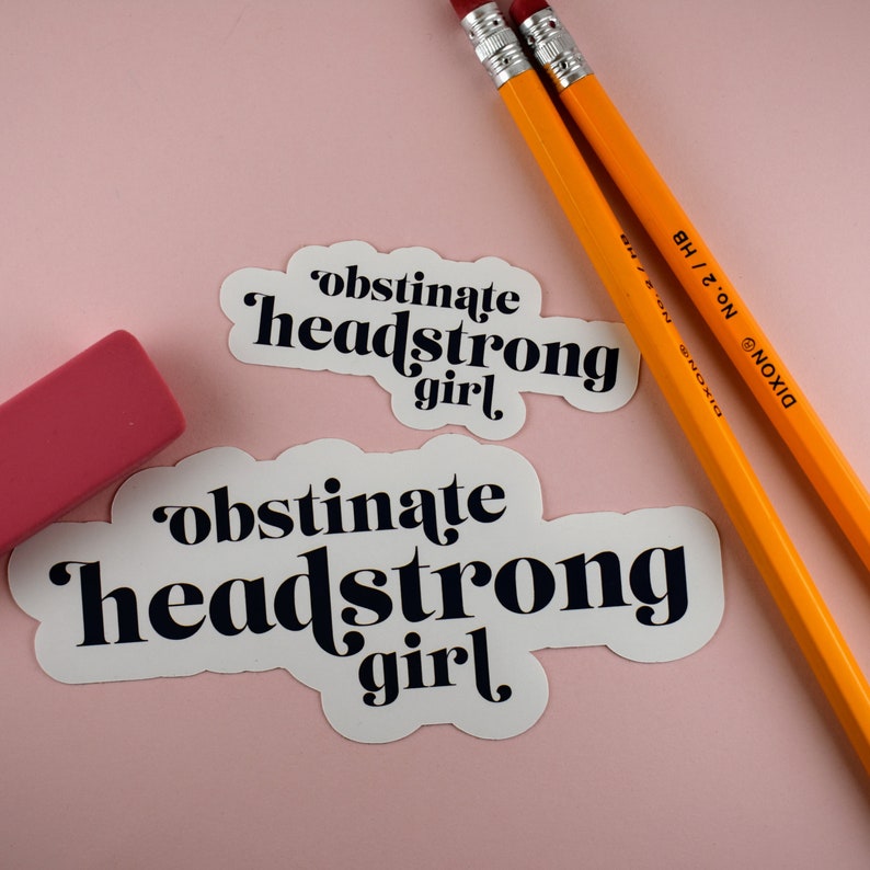 Op de afbeelding: Twee witte stickers met zwarte tekst die "obstinate headstrong girl" luidt, op een roze achtergrond. Twee gele potloden liggen op de achtergrond.