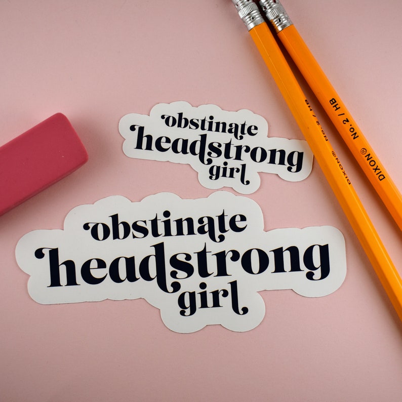 Op de afbeelding: Twee witte stickers met zwarte tekst die "obstinate headstrong girl" op een roze achtergrond zegt. De stickers zijn naast twee gele potloden geplaatst.