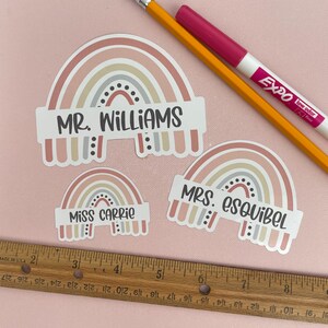 May include: Three rainbow-themed name tags with the names "MR. WILLIAMS", "MISS CARRIE", and "MRS. ESQUIBEL" printed on them. The rainbows are in shades of pink, beige, and gray.