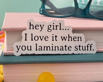 May include: White sticker with the text "hey girl... I love it when you laminate stuff." The sticker is placed on top of a stack of two books, one pink and one teal. The books are on a surface with colorful candies.