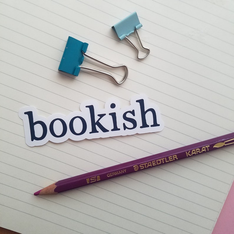 May include: A white sticker with the word "bookish" printed in dark blue letters. The sticker is on a lined notebook page with a purple pencil and two blue paper clips.