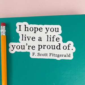 May include: A white sticker with the quote "I hope you live a life you're proud of." by F. Scott Fitzgerald. The sticker is on a teal surface, with a yellow pencil with a pink eraser in the image.
