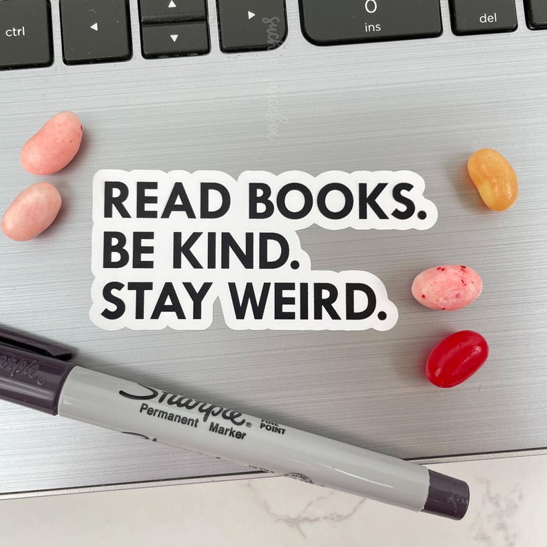 May include: A white sticker with black text that reads "READ BOOKS. BE KIND. STAY WEIRD." The sticker is on a silver laptop keyboard. There are several jelly beans scattered around the sticker.