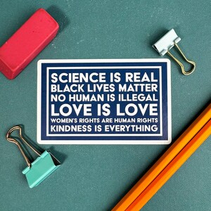 May include: A blue and white sign with the text "SCIENCE IS REAL, BLACK LIVES MATTER, NO HUMAN IS ILLEGAL, LOVE IS LOVE, WOMEN'S RIGHTS ARE HUMAN RIGHTS, KINDNESS IS EVERYTHING".