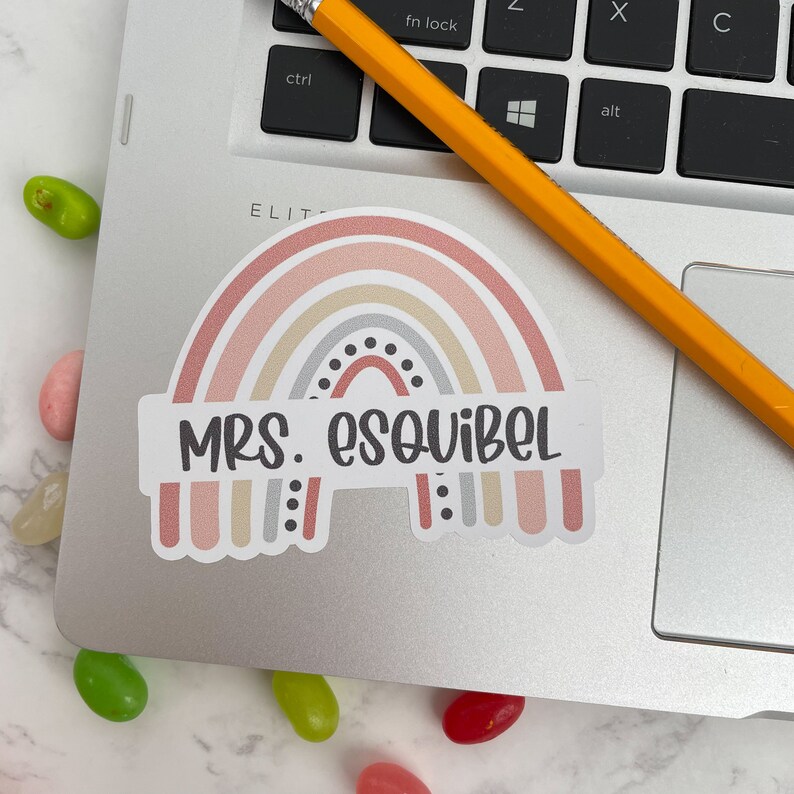 May include: A rainbow-colored sticker with a white background and the text "MRS. ESQUIBEL" in black. The rainbow has a gray center with black dots.