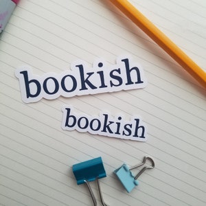 May include: Two white stickers with the word "bookish" printed in black on a lined notebook page. Two blue paper clips are on the page.