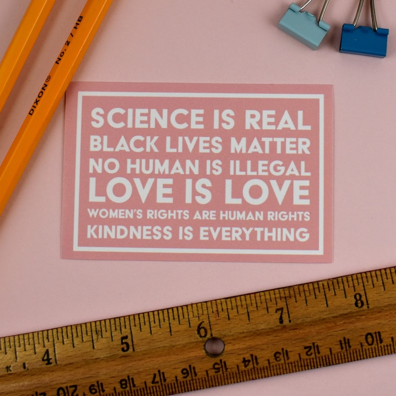 May include: A pink card with the text "SCIENCE IS REAL, BLACK LIVES MATTER, NO HUMAN IS ILLEGAL, LOVE IS LOVE, WOMEN'S RIGHTS ARE HUMAN RIGHTS, KINDNESS IS EVERYTHING" printed in white.