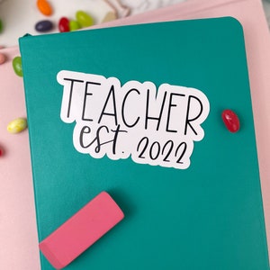May include: A teal notebook with a white sticker that says "Teacher est. 2022".