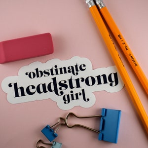 Op de afbeelding: Witte sticker met de tekst "obstinate headstrong girl" in zwarte letters. De sticker zit op een roze achtergrond met twee gele potloden en een roze gum.