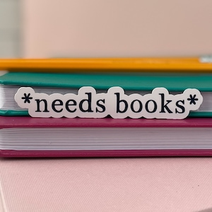 May include: A white sticker with the text "*needs books*" in black lettering, placed on top of a stack of three books. The top book is yellow, the middle book is teal, and the bottom book is pink.