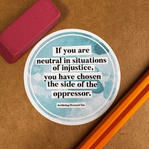 May include: A round sticker with a blue watercolor background and white text that reads "If you are neutral in situations of injustice, you have chosen the side of the oppressor. - Archbishop Desmond Tutu".