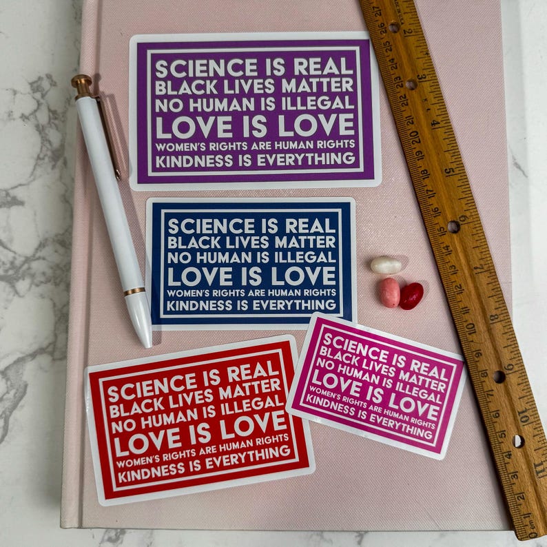 May include: Four stickers with the text "Science is real, Black Lives Matter, No Human is Illegal, Love is Love, Women's Rights are Human Rights, Kindness is Everything" in white text on a variety of colored backgrounds. The stickers are pink, blue, red, and white.