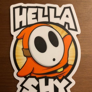 Puede incluir: Una pegatina con un personaje de dibujos animados con una máscara blanca y un cuerpo naranja. El personaje está rodeado de un sol amarillo. El texto "Hella Shy" está escrito en letras blancas y negras.