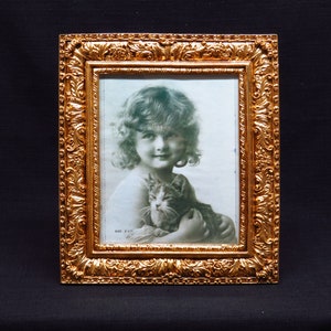 May include: An antique gold-coloured picture frame with a portrait of a child and a cat. The frame has detailed scrollwork and a rectangular shape. The portrait is in black and white, with the text "SIZE 8 x 11" visible in the bottom corner.
