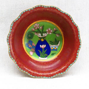 May include: A small, red, round bowl with a scalloped edge. The bowl is decorated with a green, blue, and gold floral design. The design features a vase with flowers and leaves.