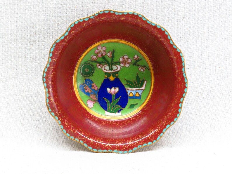 May include: A small, red, round bowl with a scalloped edge. The bowl is decorated with a green, blue, and gold floral design. The design features a vase with flowers and leaves.