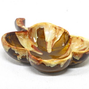 Könnte beinhalten: Eine braune und gelbe Keramikschale in Form eines vierblättrigen Kleeblatts mit einem Griff. Die Schale hat eine glasierte Oberfläche mit braunen, gelben und weißen Streifen.