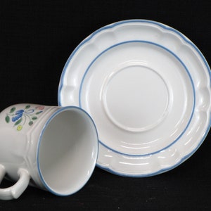 May include: A white ceramic teacup and saucer with a blue and floral design. The cup has a handle and the saucer has a scalloped edge.