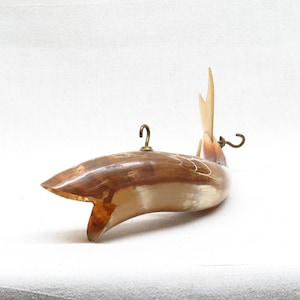 K&ouml;nnte beinhalten: Ein geschnitzter Holzfisch mit hellbrauner und wei&szlig;er Oberfl&auml;che. Der Fisch hat einen Messinghaken oben und einen Messinghaken am Schwanz. Der Fisch h&auml;ngt vor einem wei&szlig;en Hintergrund.