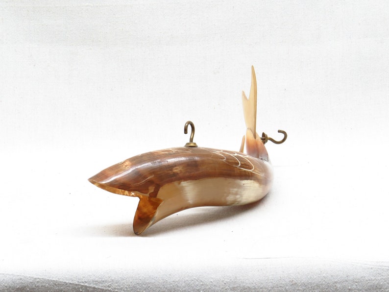 K&ouml;nnte beinhalten: Ein geschnitzter Holzfisch mit hellbrauner und wei&szlig;er Oberfl&auml;che. Der Fisch hat einen Messinghaken oben und einen Messinghaken am Schwanz. Der Fisch h&auml;ngt vor einem wei&szlig;en Hintergrund.