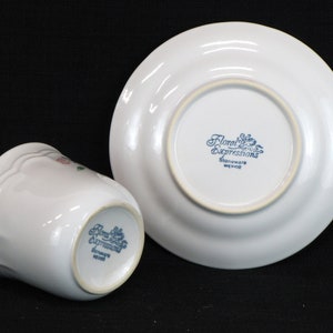 May include: A white ceramic cup and saucer with a floral design. The bottom of the cup and saucer have the words "Floral Expressions Stoneware Mexico" printed in blue.