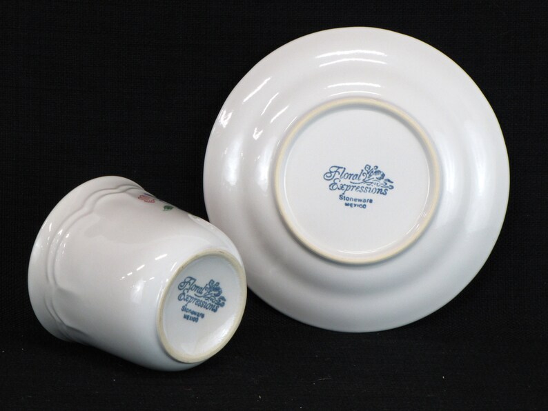 May include: A white ceramic cup and saucer with a floral design. The bottom of the cup and saucer have the words "Floral Expressions Stoneware Mexico" printed in blue.