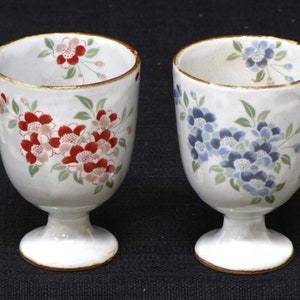 Puede incluir: Dos tazas de cerámica blanca con diseños florales. Una taza tiene flores rojas y rosas, la otra tiene flores azules. Ambas tazas tienen un borde dorado y una base de pedestal.