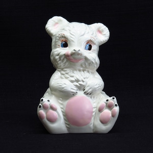 May include: White ceramic bear figurine with pink accents and blue eyes. The bear is sitting with its paws in front of it and has a pink belly.