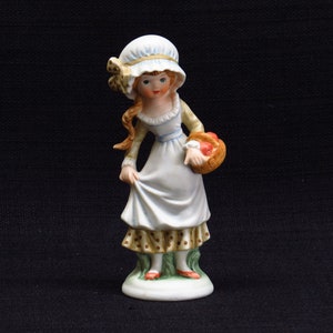 Pode incluir: Uma figura de porcelana de uma menina vestindo um vestido branco com bolinhas marrons e um chapéu azul e branco. Ela está segurando uma cesta de maçãs vermelhas.